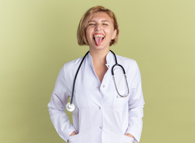 Joyful with closed eyes young female doctor wearing medical robe with stethoscope showing tongue isolated on olive green wall