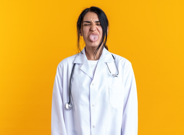 노란색 벽에 고립 된 혀를 보여주는 청진 기 의료 가운을 입고 닫힌 눈 젊은 여성 의사와 즐거운