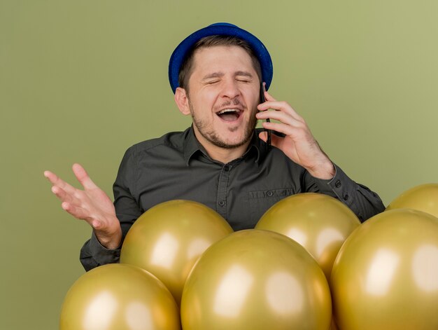 Joyful with closed eyes young party guy wearing black shirt and blue hat standing behind balloons speaks on phone and spreading hand isolated on olive green