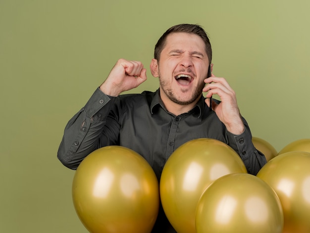 Joyful with closed eyes young party guy wearing black shirt standing among balloons speaks on phone and showing yes gesture isolated on olive green