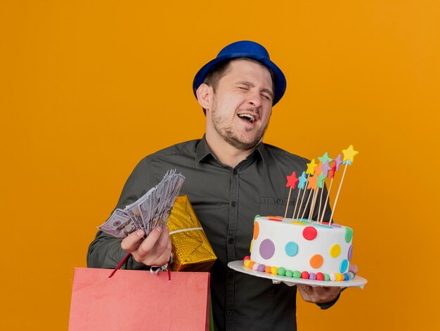 Joyful with closed eyes young party guy wearing blue hat holding cake with gifts and cash isolated on orange