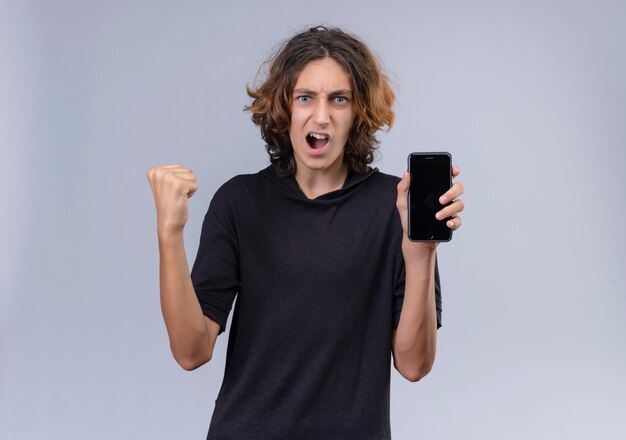 Радостный с эмоциями парень с длинными волосами в черной футболке держит телефон на белой стене