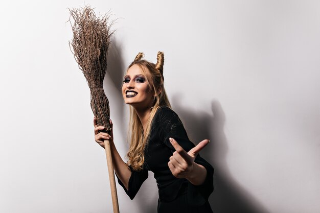 Joyful wizard in big hat having fun in halloween. Pretty young witch with broom dancing with smile.