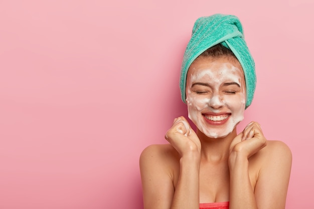 Free photo joyful woman clenches fists, smiles broadly, shows white teeth, washes face with soap, removes dirt, enjoys hygienic treatments at home, wrapped in soft towel