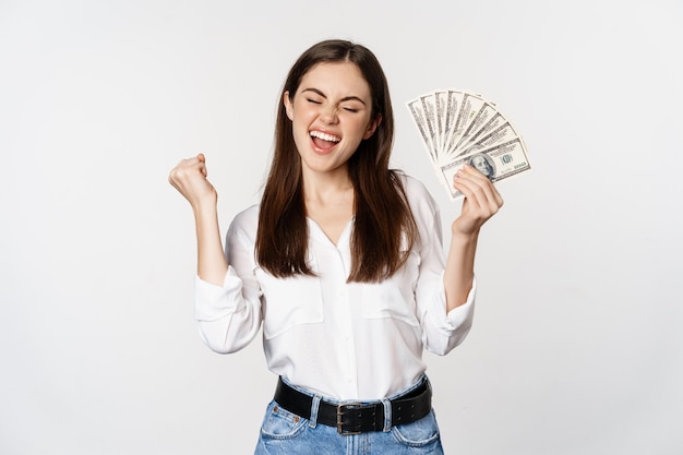 Joyful woman dancing with money smiling pleased winning prize standing over white background