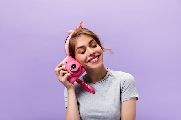 Бесплатное фото Радостная женщина счастливо позирует с розовой камерой. довольно яркая девушка в серой футболке и летней повязке на голову, улыбаясь на фиолетовом фоне.