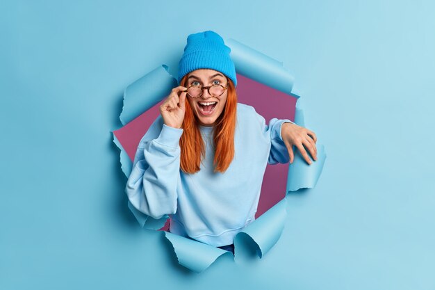 Joyful woman has red hair smiles broadly has happy curious expression keeps hand on eyeglasses laughs positively dressed in blue clothes breaks through paper hole