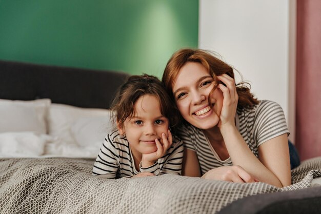 Радостная женщина и ее милая дочь лежат на кровати и с улыбкой смотрят в камеру