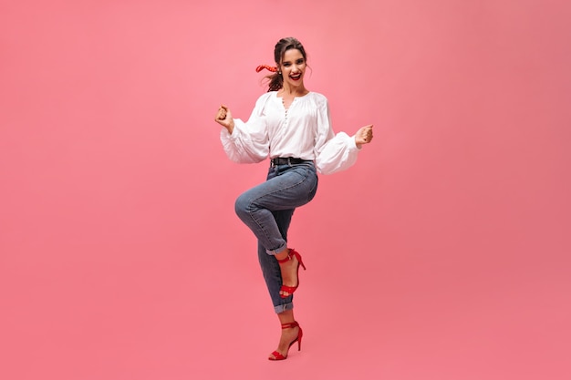 Joyful woman in jeans, white blouse dancing on pink background.  Modern girl with red lipstick and in stylish heels rejoices on isolated backdrop.