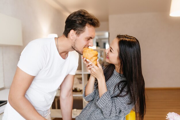 Joyful woman in knitted woolen sweater feeding husband with sweet croissant and laughing