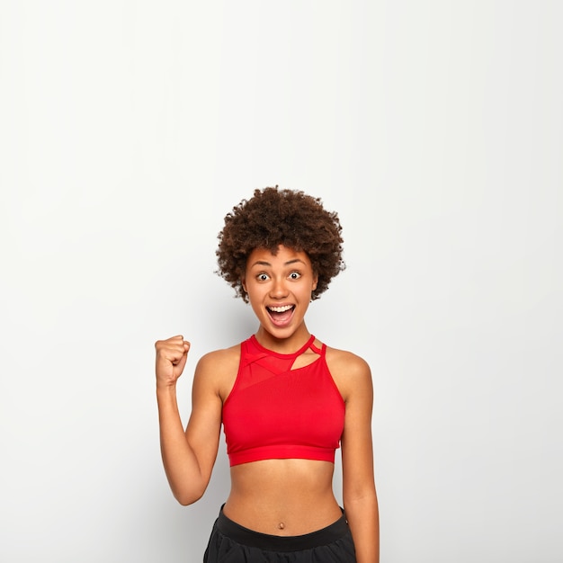 Joyful woman raises clenched fist, cheers for something, gestures with triumph, has perfect figure, wears red top, poses against white background, blank space above