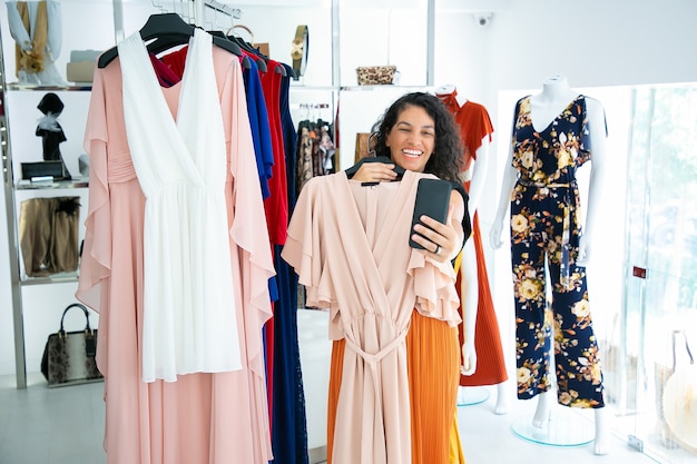 Радостная женщина делает покупки в магазине одежды и консультирует друга по мобильному телефону, показывая платье на вешалке. Бутик-клиент или концепция коммуникации