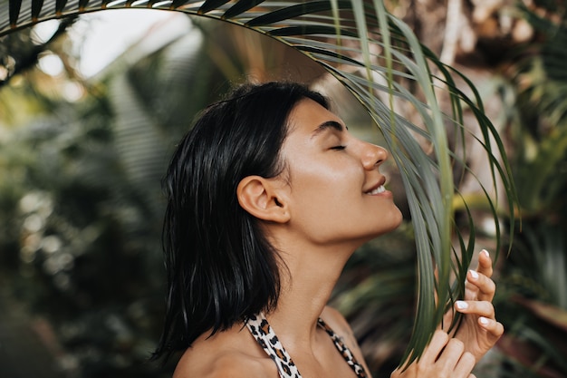 Joyful woman sniffing palm tree with closed eyes. Outdoor shot of beautiful tanned woman enjoying vacation.