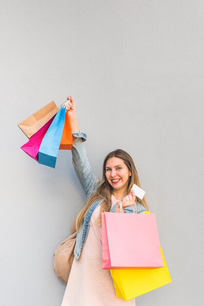 Joyful woman standing with shopping bags and credit card at light wall