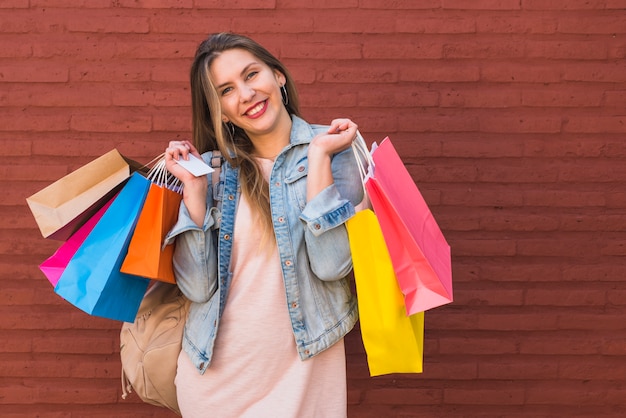 Joyful woman standing with shopping bags and credit card at red brick wall