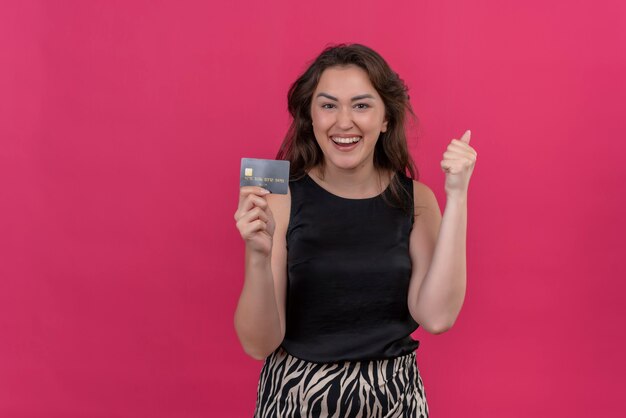 Joyful woman wearing black undershirt holding a credit card on pink wall