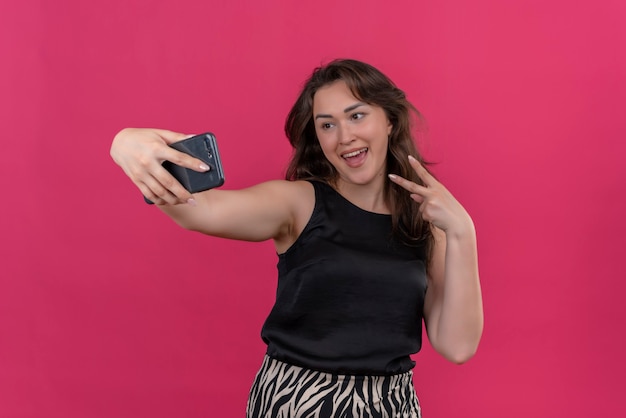 Free photo joyful woman wearing black undershirt take a selfie and show viva gesture on pink wall