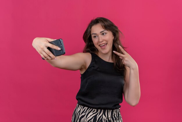 Joyful woman wearing black undershirt take a selfie and show viva gesture on pink wall