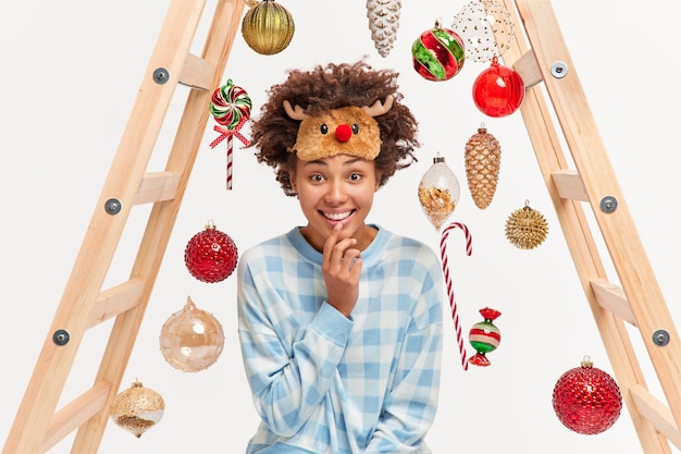 Joyful woman wears domestic pajama and sleep mask looks with happy smile at camera poses 