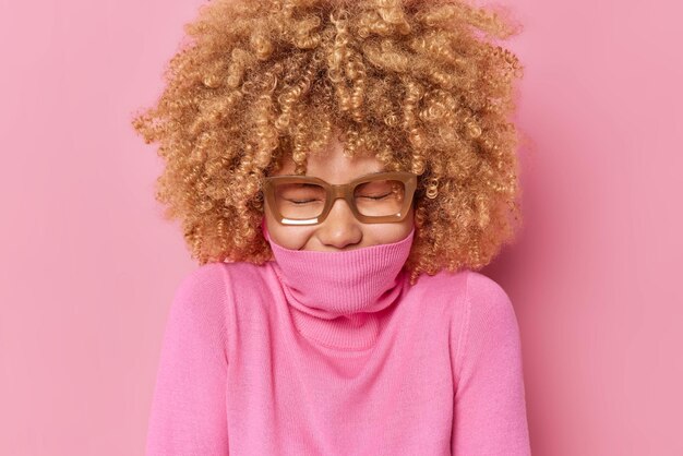 Joyful woman with bushy curly hair expresses sincere emotions keeps eyes closed covers mouth with collar has upbeat mood isolated over pink background hides face. Human sincere feelings concept