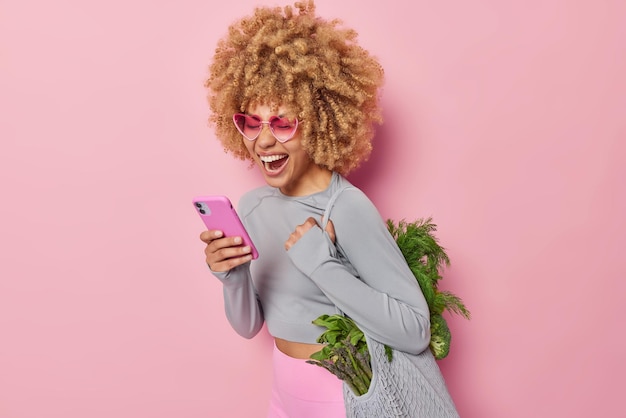 巻き毛のふさふさした髪のうれしそうな女性は、携帯電話を使用してオンラインショッピングを行い、ピンクの背景で隔離されたベジタリアンサラダを作るための健康的な緑の野菜でいっぱいのファブリックバッグを運びます