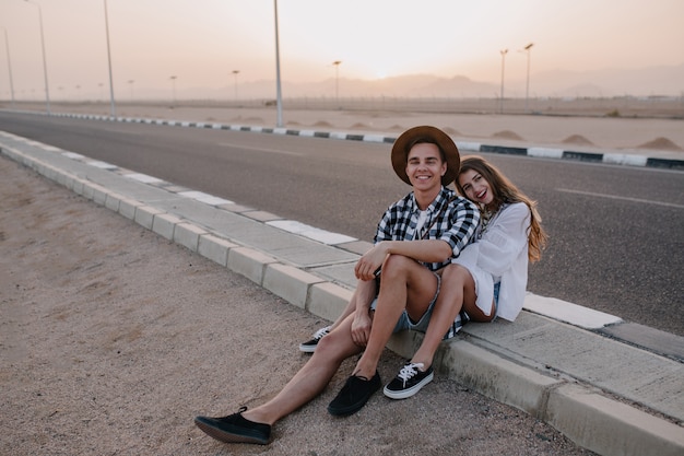 Donna allegra con acconciatura carina seduta sulla strada, rannicchiata contro il suo fidanzato in cappello alla moda e ridendo. affascinante giovane donna e uomo che riposa vicino all'autostrada dopo il viaggio e gode del tramonto.