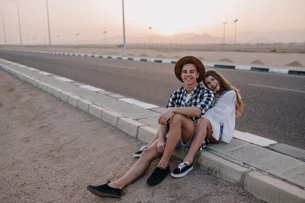 Joyful woman with cute hairstyle sitting on the road, huddled against her boyfriend in trendy hat and laughing. Charming young woman and man resting near the highway after travel and enjoys sunset.