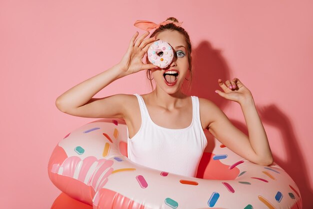 Joyful woman with gold earrings in modern white swimsuit holding donut and posing with large swimming suit on pink background