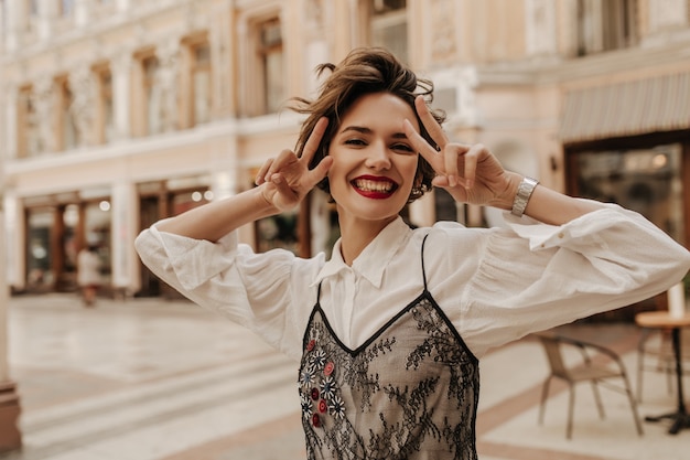 Joyful woman with red lips showing peace signs in city. Modern woman with short hair in white blouse with black lace smiling at street.