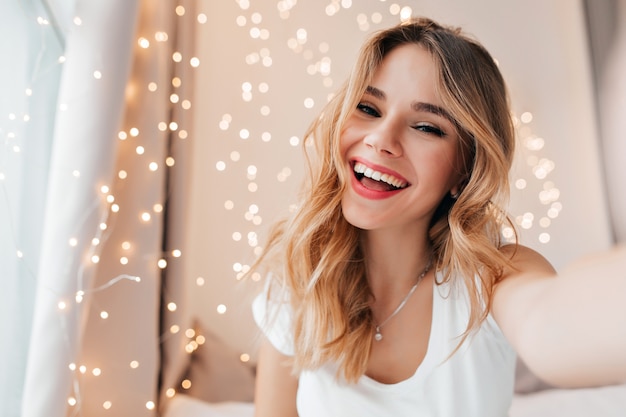 Joyful woman with sincere smile posing in her room. Blonde caucasian girl expressing happiness