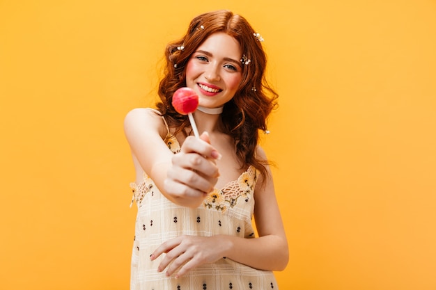黄色いサンドレスのうれしそうな女性はピンクのお菓子を持っています。オレンジ色の背景に彼女の髪に花を持つ女性の肖像画。