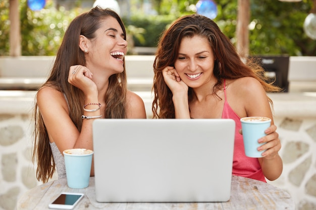 Joyful women have fun together, laugh while watch comedy online on laptop computer and recreat in outdoor cafe, drink hot coffee or espresso