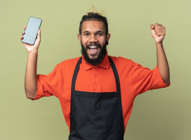 Joyful young afro-american barber wearing uniform holding mobile phone  doing yes gesture isolated on olive green wall