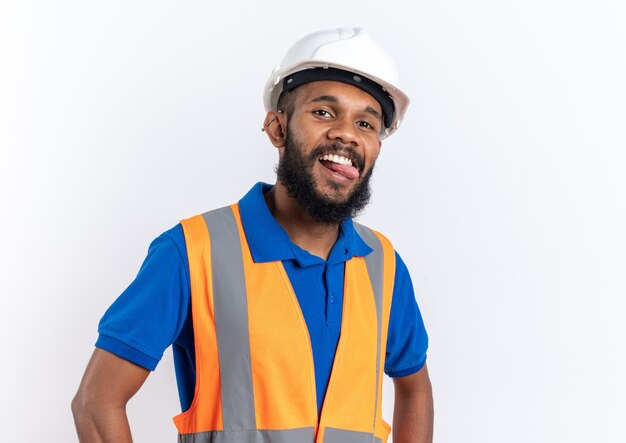 Joyful young afro-american builder man in uniform with safety helmet stucks out his tongue isolated on white background with copy space