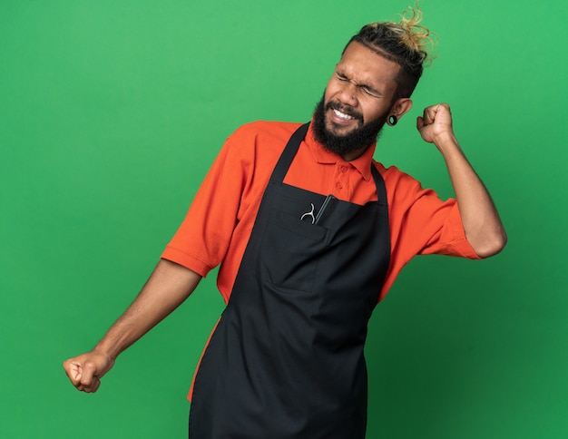 Радостный молодой афро-американский парикмахер-мужчина в униформе делает жест `` да '' с закрытыми глазами, изолированными на зеленой стене
