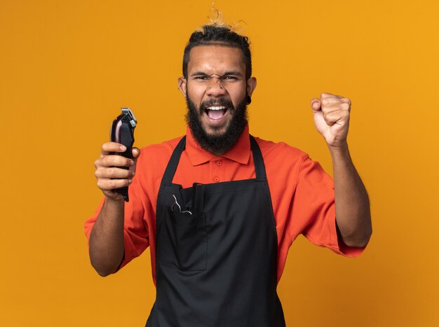 Joyful young afro-american male barber wearing uniform holding hair clippers doing yes gesture 