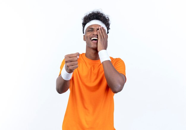 Joyful young afro-american sporty man wearing headband and wristband covered eye with hand and holding out fist to camera isolated on white background