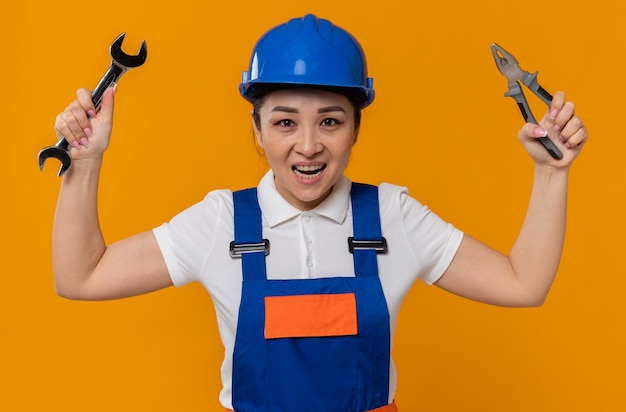 Foto gratuita gioiosa giovane ragazza asiatica del costruttore con il casco di sicurezza blu che tiene le pinze e la chiave dell'officina