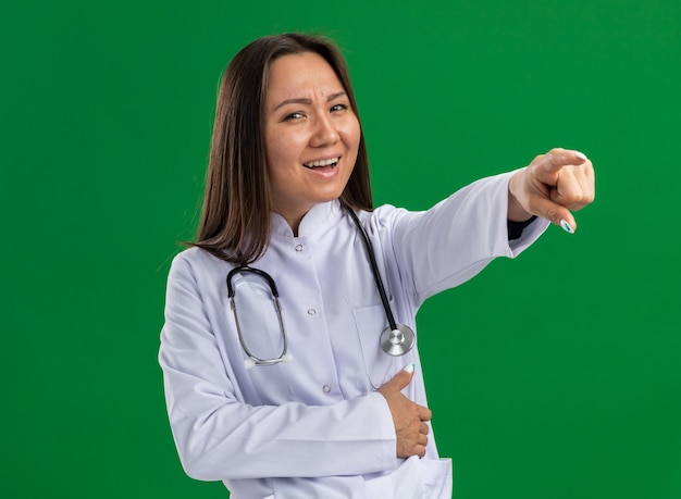 Joyful young asian female doctor wearing medical robe and stethoscope looking at camera pointing at side keeping hand on belly isolated on green wall