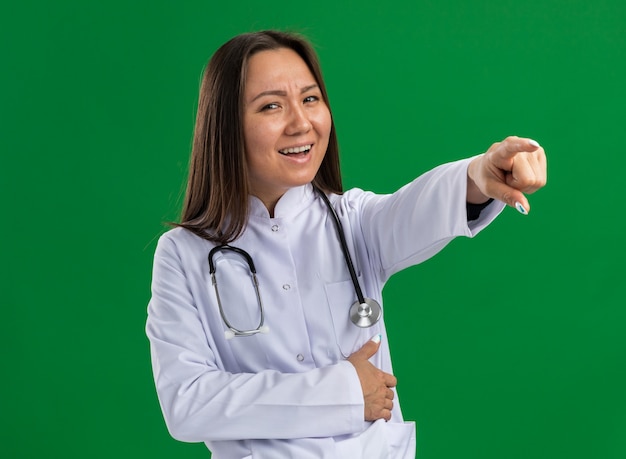 의료 가운과 청진기를 착용하고 녹색 벽에 고립 된 배에 손을 잡고 측면을 가리키는 카메라를보고 즐거운 젊은 아시아 여성 의사