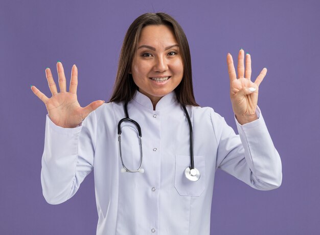 Joyful young asian female doctor wearing medical robe and stethoscope looking at front showing nine with hands isolated on purple wall