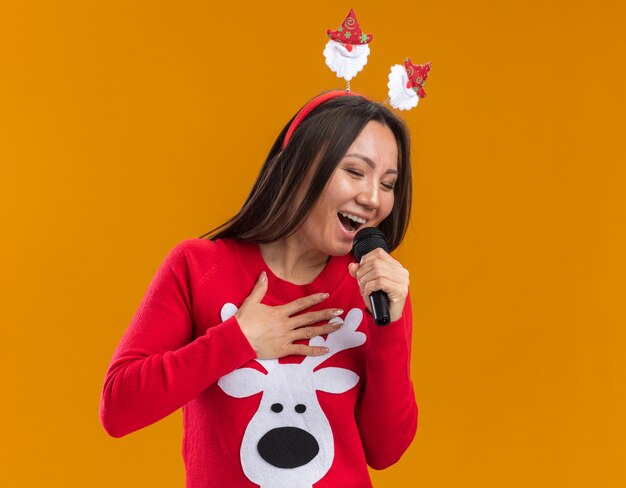 Joyful young asian girl wearing christmas hair hoop with sweater sings on microphone isolated on orange wall