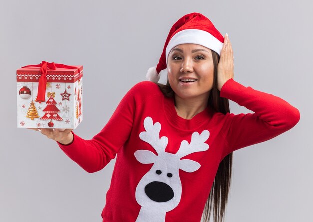 Joyful young asian girl wearing christmas hat with sweater holding gift box putting hand on cheek isolated on white background