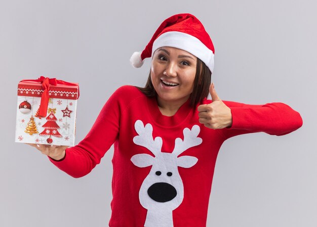 Joyful young asian girl wearing christmas hat with sweater holding gift box showing thumb up isolated on white background