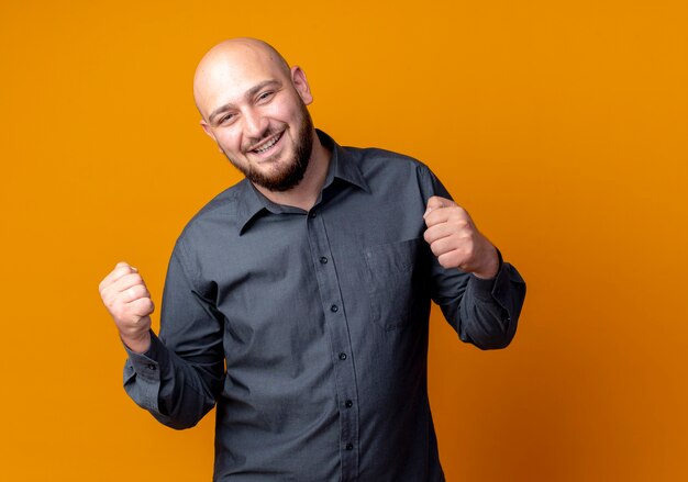 Joyful young bald call center man clenching fists isolated on orange