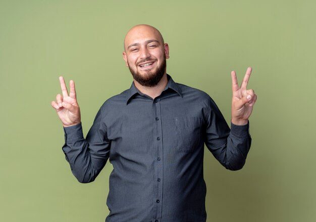 Joyful young bald call center man doing peace sign isolated on olive green