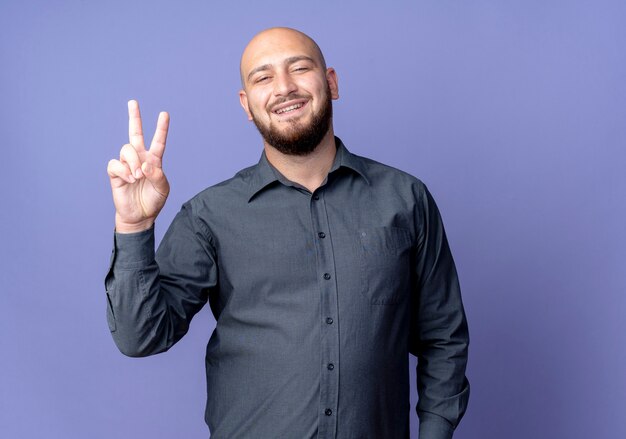 Joyful young bald call center man doing peace sign isolated on purple