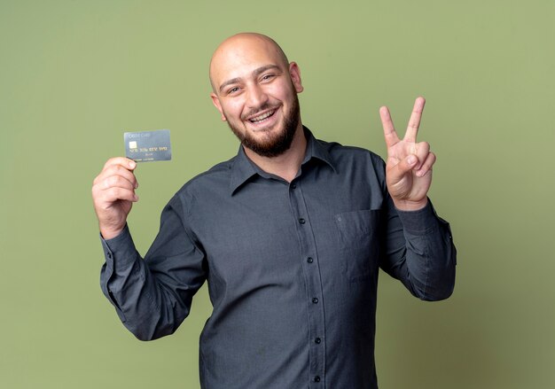 Joyful young bald call center man holding credit card doing peace sign isolated on olive green