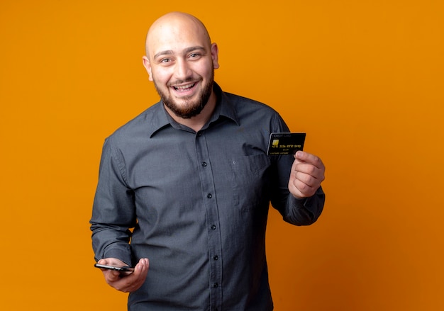 Joyful young bald call center man holding credit card and mobile phone isolated on orange