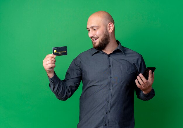 Joyful young bald call center man holding credit card and mobile phone looking at card isolated on green